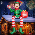 Giant elf inflatable with striped legs sitting on a snowy cabin roof holding a candy cane and a gift